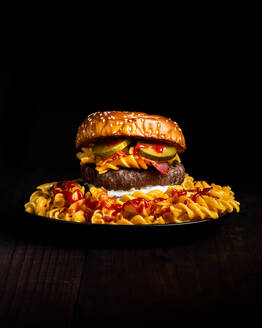 Hamburger placed on plate among macaroni ketchup and cheese against dark background - ADSF48530