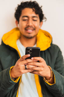 Young ethnic content unshaven male in formal apparel with cellphone and wireless earbud looking at screen on white background - ADSF48535