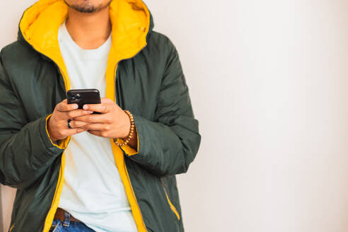 Anonymous young ethnic male in formal apparel with cellphone and wireless earbud looking at screen on white background - ADSF48539