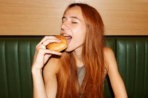 Young woman with red hair and piercing with closed eyes sitting at table and enjoy eating hamburger in restaurant - ADSF48615