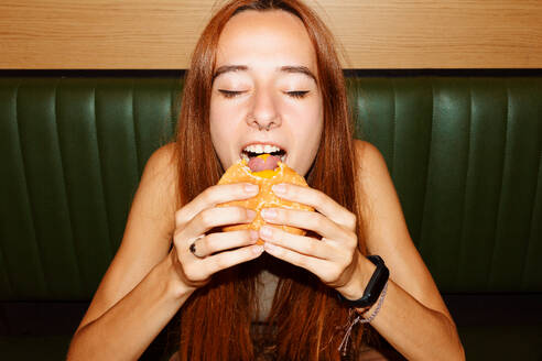 Young woman with red hair and piercing with closed eyes sitting at table and enjoy eating hamburger in restaurant - ADSF48616