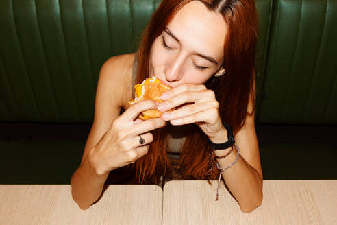 High angle of young woman with red hair and piercing with closed eyes sitting at table and enjoy eating hamburger in restaurant - ADSF48617