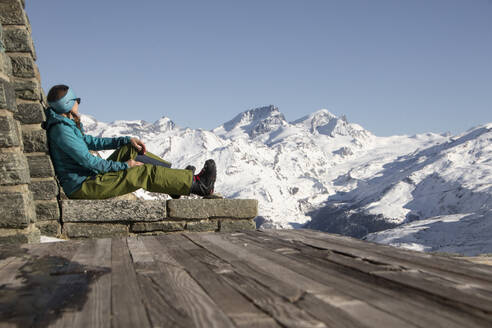 Side view of adult long hair woman sitting on snowy on a rock looking at the mountain in winter in zermatt, Switzerland - ADSF48632