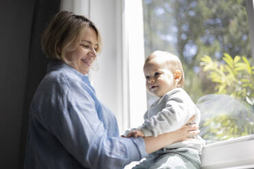 Side view of mature blond woman smiling with a baby boy near the window at home - ADSF48676