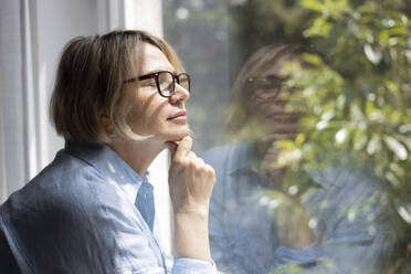 Side view of mature blond woman with eyeglasses closed eyes and hand on chin standing near the window at home - ADSF48677