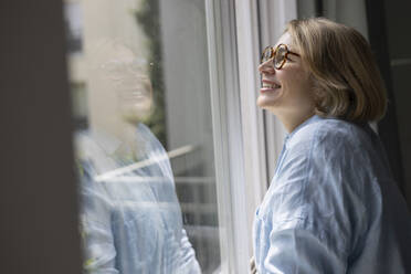 Side view of mature blond woman with eyeglasses standing and looking though the window at home - ADSF48678