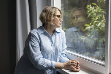 Side view of mature blond woman with eyeglasses standing and looking though the window at home - ADSF48679