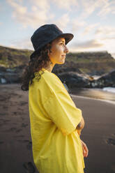 Pensive young female in casual clothes and hat standing on sandy seashore while looking away against waving sea - ADSF48684