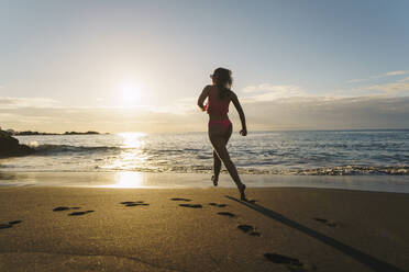 Full body of anonymous cheerful barefoot female in bikini running along seaside and on sunny day in Spain - ADSF48685