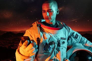 Ethnic male astronaut in spacesuit holding helmet while browsing smartphone and standing on ground over glowing space - ADSF48688