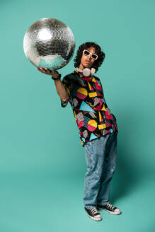 Funky young Latin American male with sunglasses looking away while standing with raised arm carrying disco ball at turquoise background - ADSF48703