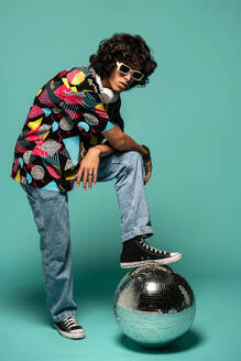 Funky young Latin American male with headphones sunglasses looking at camera while standing with leg on sphere leaning on knee at turquoise background - ADSF48704