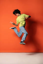 Full body side view of young Latin American male dancer looking down while jumping above ground in dance pose with victory sign against red background - ADSF48706