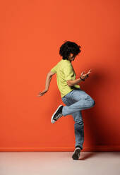 Side view of young male with curly hair while dancing in sneakers with hand and leg raised in light with shadow against red background - ADSF48707