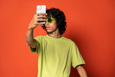 Smiling young Latin American male with dark hair looking at screen of mobile phone while standing with sunglasses during selfie at red background - ADSF48709