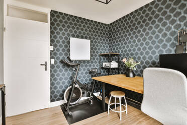 Spacious room with wooden desk and exercise bike in contemporary house with black wallpaper and white closed door - ADSF48725