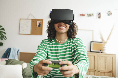 Smiling girl wearing virtual reality simulators playing video game with controller at home - ALKF00663