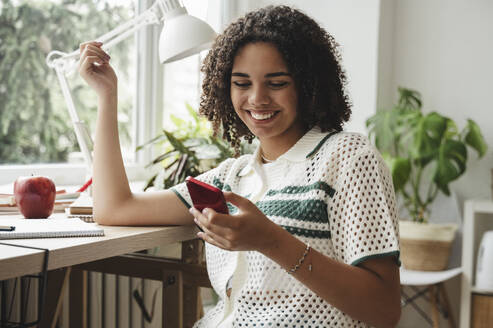 Happy curly haired girl using smart phone at desk - ALKF00695