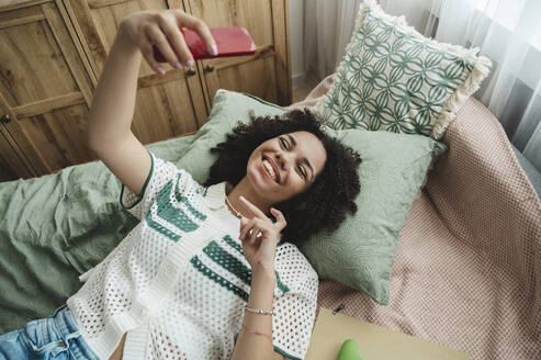 Happy teenage girl taking selfie through smart phone on bed at home - ALKF00711