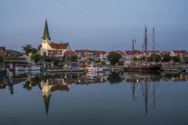 Dänemark, Bornholm, Ronne, St.-Nikolaus-Kirche und umliegende Häuser spiegeln sich in der Abenddämmerung im Küstenwasser - KEBF02767