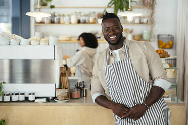 Smiling barista wearing apron standing in front of counter at coffee shop - EBSF03995