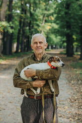 Smiling senior man holding Jack Russell Terrier dog in arms at park - VSNF01411