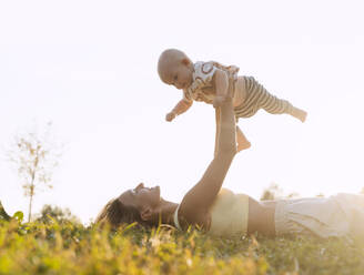 Smiling mother lying on grass and holding baby boy under sky at sunset - NDEF01283
