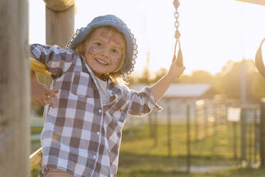 Lächelndes Mädchen mit Gesichtsbemalung spielt auf einem Spielplatz bei Sonnenuntergang - NDEF01289