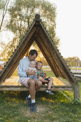 Liebevoller Vater sitzt mit Kindern in einer dreieckigen Holzhütte im Park - NDEF01291