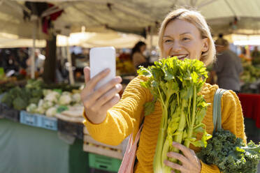 Woman taking selfie with celery through smart phone at farmer's market - NDEF01308
