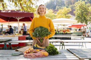 Smiling woman holding green leafy vegetables standing near table at farmer's market - NDEF01311