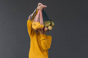 Frau bedeckt Gesicht mit Früchten in Netztaschen vor schwarzem Hintergrund - NDEF01320