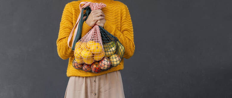 Woman holding fresh fruits in mesh bags against black background - NDEF01321