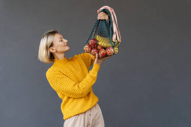 Lächelnde Frau schaut auf Netztaschen mit Früchten vor schwarzem Hintergrund - NDEF01322