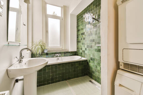 Ceramic washbasin and bathtub with green tiled walls by window in spacious bathroom at modern apartment - ADSF48774