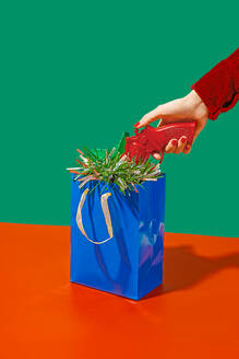 Crop anonymous woman near blue carton bag placed on red surface with tinsel taking red toy gun against green background - ADSF48788