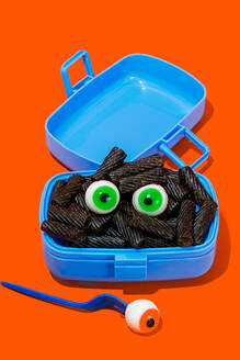 Horror lunch with black pasta and eyes in lunchbox placed on orange background near fork with eye - ADSF48790