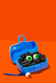 Horror lunch with black pasta and eyes in lunchbox placed on orange background near fork with eye - ADSF48791