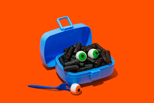 Horror lunch with black pasta and eyes in lunchbox placed on orange background near fork with eye - ADSF48792