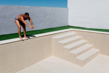 Side view of strong male athlete in sportswear looking down while standing on edge of empty swimming pool with stairs pretending to jump in daylight - ADSF48798