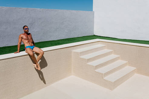 Muscular male in swimwear sitting on poolside of empty swimming pool with staircase against cloudless blue sky in day - ADSF48799