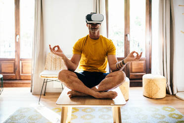 Full body of anonymous male in VR goggles while sitting in room on table in Padmasana pose with Gyan mudra and practicing yoga in cyberspace in day - ADSF48801