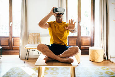 Full body of bearded male adjusting VR goggles with crossed legs and stretching arm while training in light apartment - ADSF48802