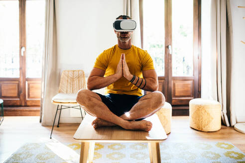 Full body of bearded male in VR goggles with crossed legs while practicing yoga in light apartment - ADSF48803