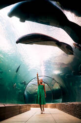 Entzückendes Mädchen in lässigem grünem Kleid, das in einem Glastunnel unter einem Aquarium steht und auf ein schwimmendes Paar Seekühe in hellem, welligem Wasser mit Fischen zeigt - ADSF48807