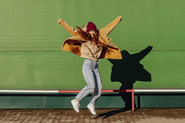 Smiling woman with arms raised jumping in front of green wall - ADF00209