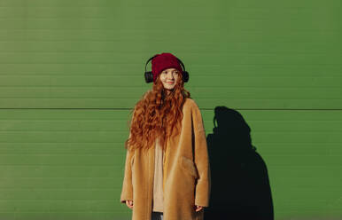 Smiling woman wearing wireless headphones standing in front of green wall - ADF00212