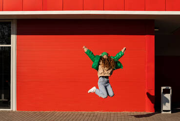Junge Frau mit erhobenen Armen, die vor eine rote Wand springt - ADF00214