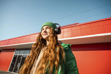 Lächelnde Frau mit langen Haaren und drahtlosen Kopfhörern vor einem Gebäude stehend - ADF00216