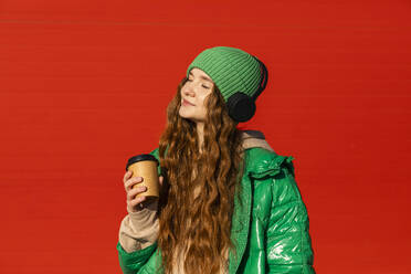 Glückliche Frau mit drahtlosen Kopfhörern, die eine Kaffeetasse vor einer roten Wand hält - ADF00218